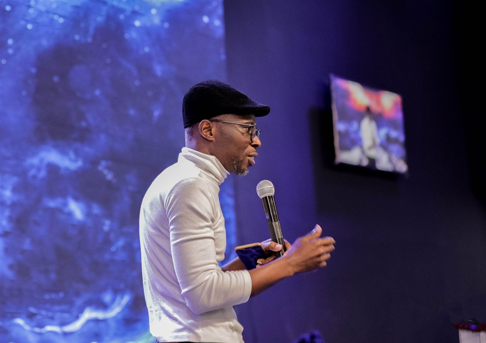 Bernard speaking on stage in a white top and black cap